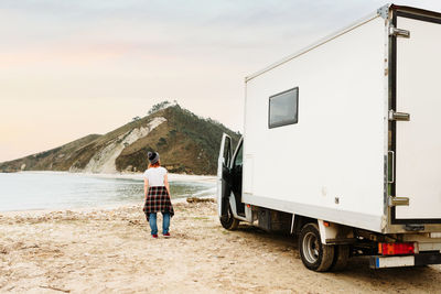 Back view of anonymous traveling female hipster standing near parked truck on shore of lake in mountainous area