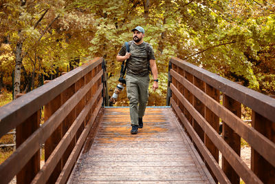 Wildlife photographer in camouflage clothing crosses a wooden bridge in the forest.