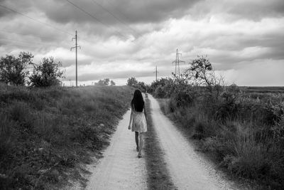 Rear view of woman walking on dirt road amidst plants against cloudy sky