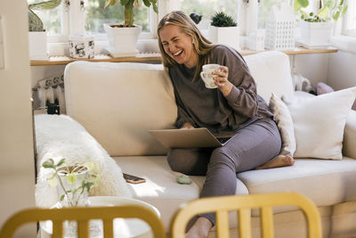 Happy female freelancer with laptop holding mug while sitting on sofa at home