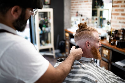 Side view modern hairdresser barber cutting a adult man redhead's hair in barber chair