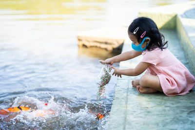 Cute girl wearing protective face mask feeding carp koi fishes at pond.