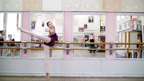 In dancing hall, young ballerina in purple leotard performs developpe attitude on pointe shoes