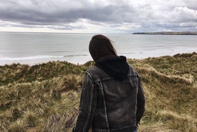 Rear view of young man looking at sea while standing against cloudy sky