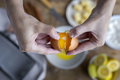 From above top view unrecognizable female breaking fresh chicken egg into bowl while cooking pastry in a wooden table with fresh ingredients