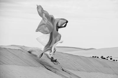 Woman with fabric jumping over sand at beach against sky