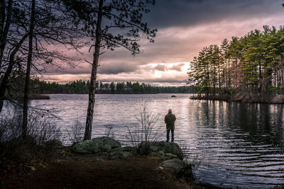 Rear view of man standing by lake against cloudy sky at sunset