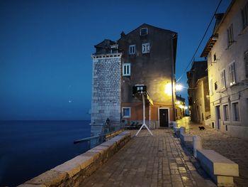 Illuminated street by buildings against sky at night