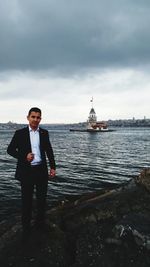 Portrait of young man standing at sea shore against cloudy sky