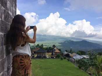 Rear view of woman photographing of landscape against cloudy sky with mobile phone