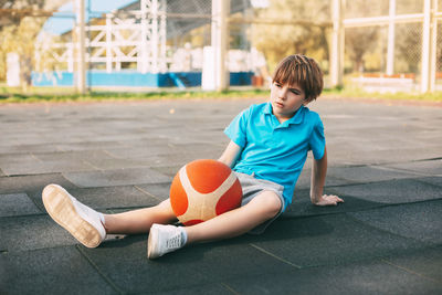 A boy basketball player in a blue shirt is sitting on the basketball court with a ball in his hands