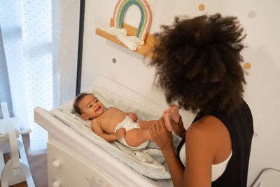 From above of tender african american mother caressing adorable toddler lying in diaper on changing table at home