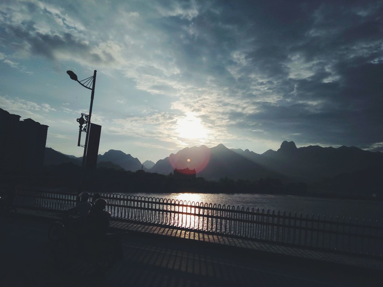 water, silhouette, mountain, tranquil scene, lake, sun, scenics, railing, tranquility, sunlight, sunset, sunbeam, mountain range, beauty in nature, calm, nature, river, outdoors, cloud - sky, majestic, day, sky, bright, lens flare, remote, no people, riverbank