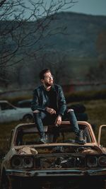 Full length of man sitting on abandoned car during dusk