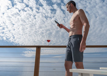 Low angle view of shirtless mature man using phone against cloudy sky