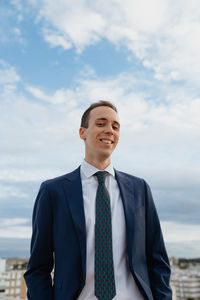 Young businessman in a suit and tie looking at camera and smiling while standing  with a cloudy sky