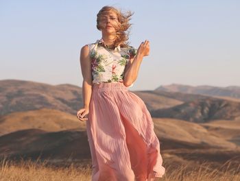 Portrait of beautiful woman in dress with tousled hair standing on field against clear sky during sunset