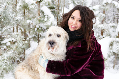 Young smiling woman is hugging her dog on a background of winter coniferous forest.