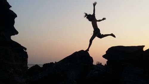 Silhouette man jumping on rock against sky at sunset