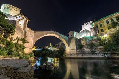 Bridge over river by illuminated buildings against sky at night
