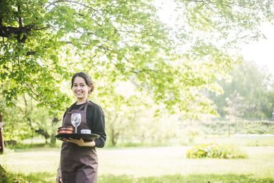 Portrait of confident young female waitress smiling while holding serving tray against plants