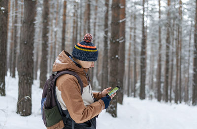 Young man with backpack looking at map on smartphone while hiking in winter snowy forest technology