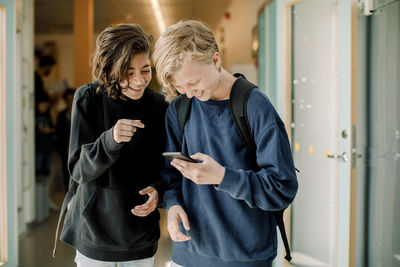 Smiling male students using smart phone in school corridor