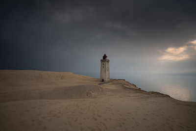 Lighthouse on beach by sea against cloudy sky