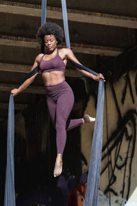 Slender african american female balancing on hanging hammock while doing aerial yoga above ground under bridge in city