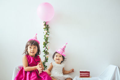 Sisters wearing party hats sitting against wall at home