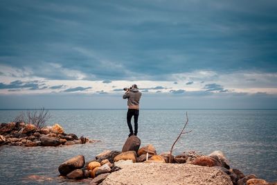 Rear view of man photographing sea while standing on rock against cloudy sky