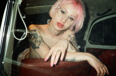 Portrait of topless sensuous tattooed woman in car seen through window at night