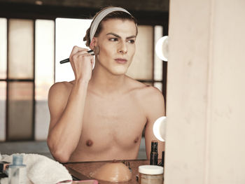 Shirtless androgynous young man with headband applying foundation on cheek while sitting in front of mirror in studio