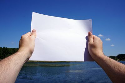 Cropped hands holding blank paper against sky