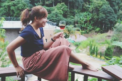 Side view of smiling mid adult woman holding wineglass while sitting on bench against trees