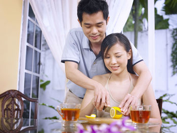 Man assisting woman in cutting fruit while having tea on glass table at home