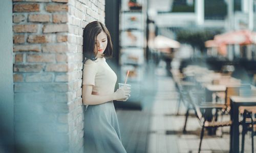 Side view of young woman holding coffee cup while standing by brick wall in city