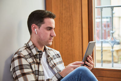 Young man seated at home using digital tablet. chilling next to the window consuming digital content.