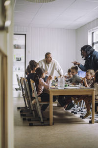 Female and male teachers talking with children having breakfast at dining table