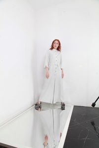 Full length fashion catalog style portrait of woman in jumpsuit standing on mirror over white wall