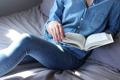 Midsection of woman reading book while sitting on bed at home