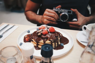 Midsection of man with camera sitting by pancake in plate on table at restaurant 