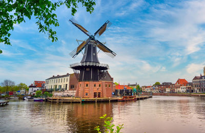 Traditional windmill by river and buildings against sky