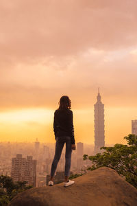 Rear view of man standing by buildings against sky during sunset