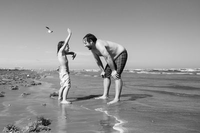 Full length of shirtless man with son on beach against sky
