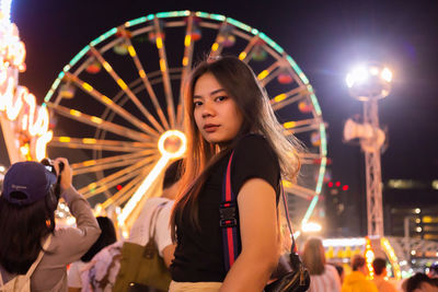 Portrait of young woman with illuminated carousel at amusement park
