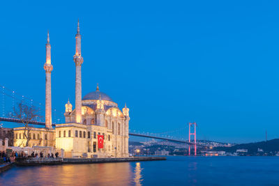 View of illuminated buildings by river against blue sky