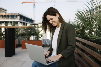 Smiling female entrepreneur using laptop while sitting on bench at building terrace