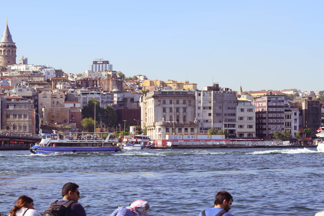 PEOPLE ON SEA BY BUILDINGS AGAINST SKY