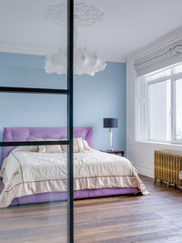 A large purple bed in a blue room near a large window. in the foreground is a glass partition